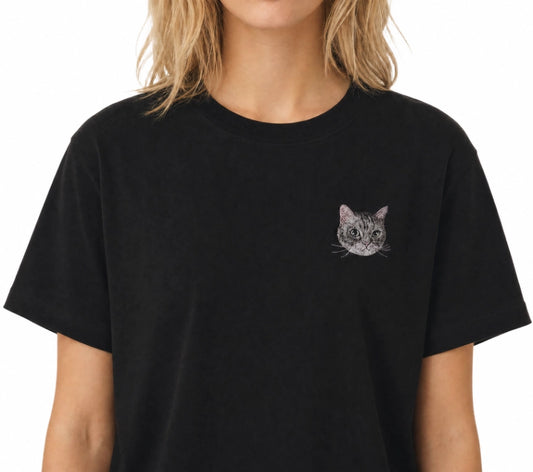 A woman wearing a black Petailor custom t-shirt with a detailed gray cat face portrait embroidered on the chest. Professional-grade realistic pet embroidery on high-quality cotton tee, perfect personalized gift for cat moms.